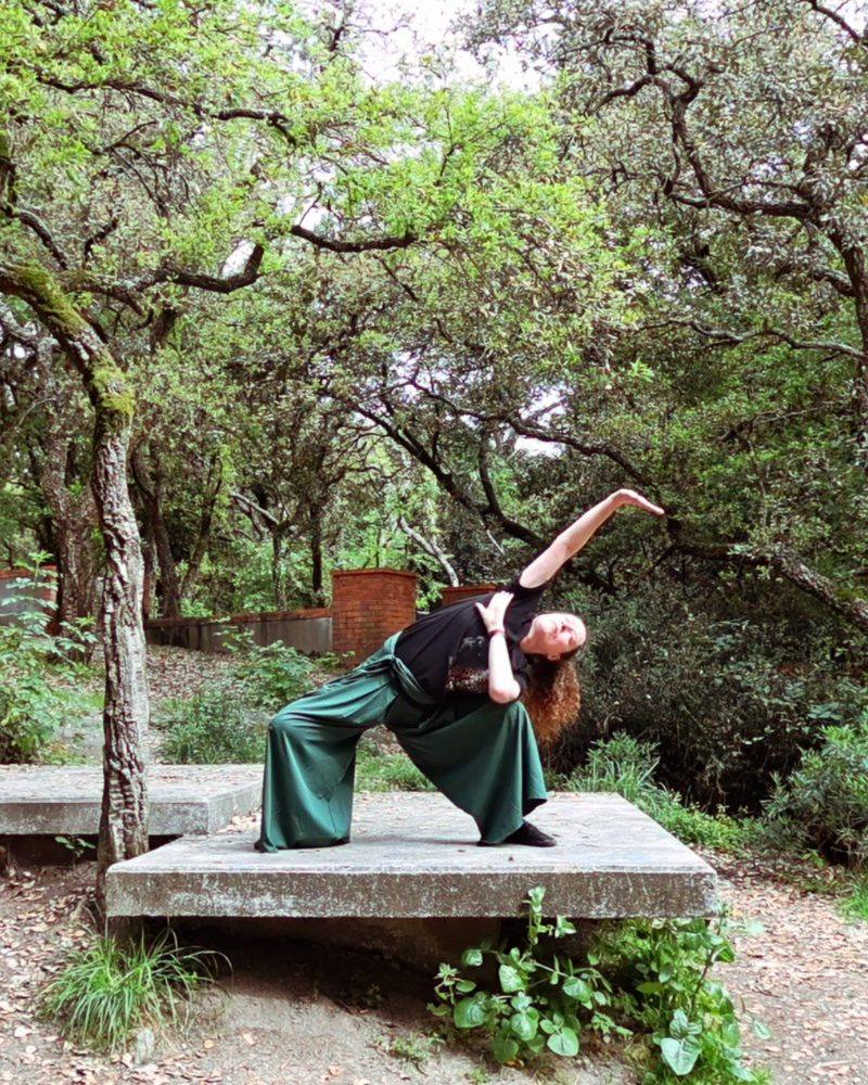 Hanna Sahlman practising Qigong outdoors, in a deep side-bending posture on a stone platform among trees.